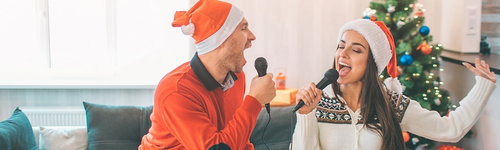 Titelbild Weihnachtslieder singen: Mann und Frau mit Weihnachtsmützen singen in Mikrofone, Weihnachtsbaum im Hintergrund